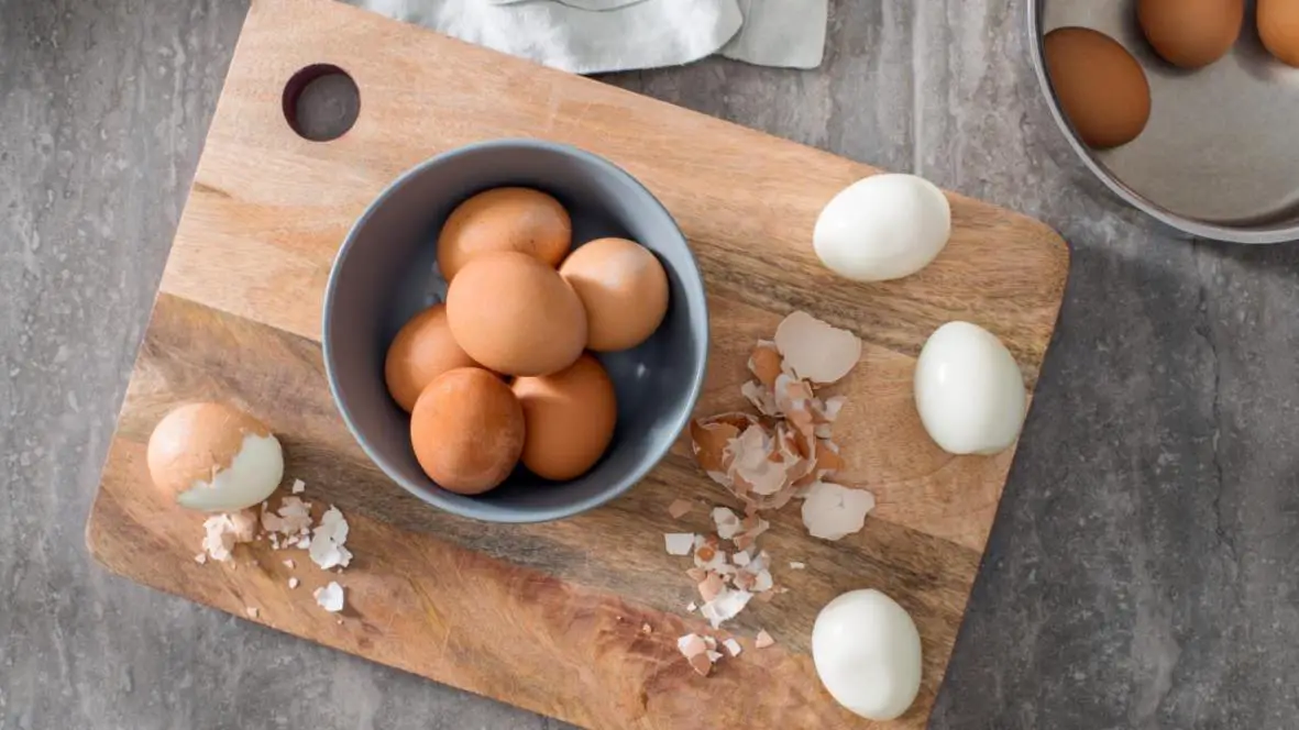 Peeled boiled eggs placed on a chopping board on a center on which bowl of unpeeled eggs is present