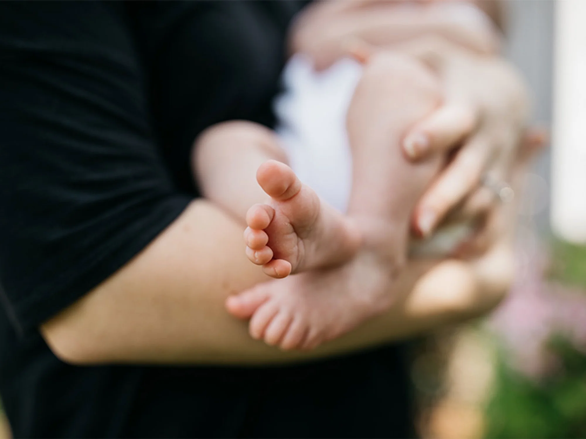 Infant's feet against unfocused background in which his mother is hold him in her arms.