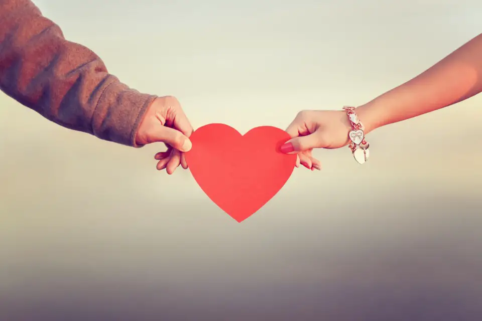 A male hand and a female hand holding red paper hearts from opposite sides