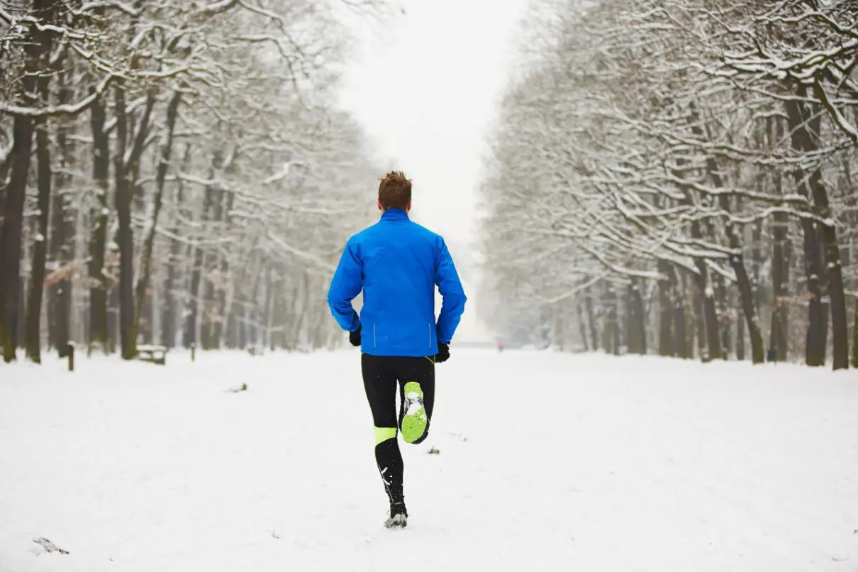 A man is running in very cold, snowy weather, where the path is covered with snow, and the trees are blanketed in a layer of snow