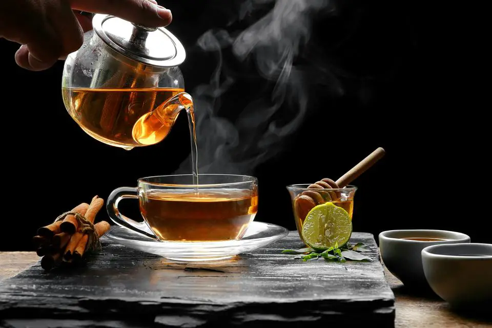 A person pours hot tea into a cup on a raised platform, surrounded by tied herbal sticks, a bowl of honey, and a side of lemon.