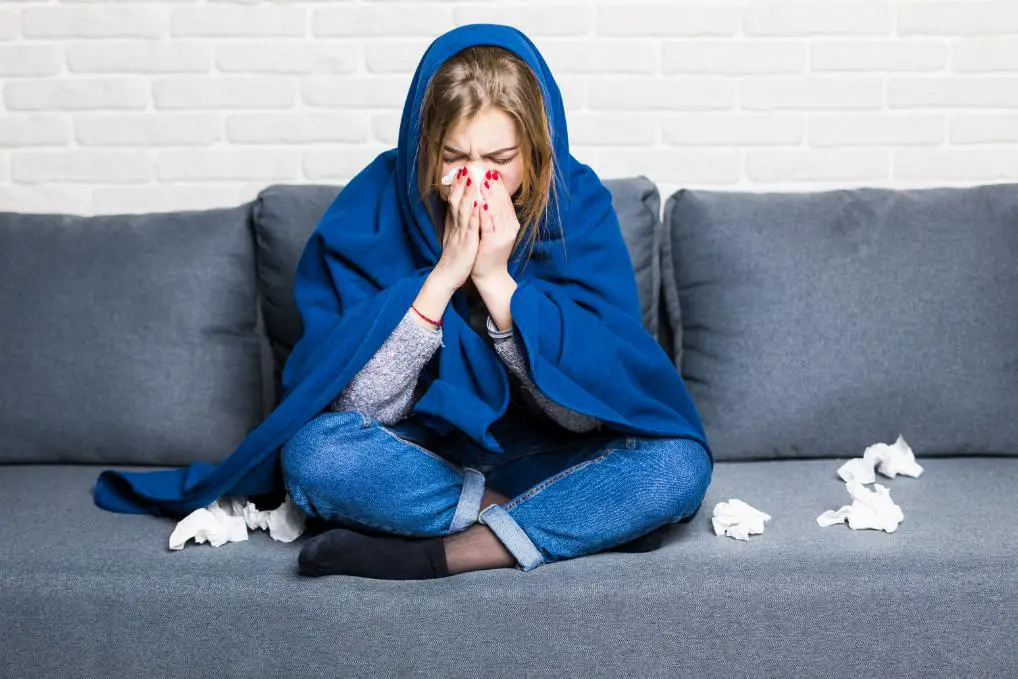 A sick girl with flu sitting on her sofa, covered with a shawl, with used tissues on the sofa
