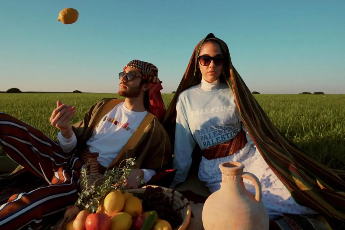 A girl and a boy are seated in a lush green field, adorned in traditional attire wearing sunglasses, and surrounded by a bounty of fruits.