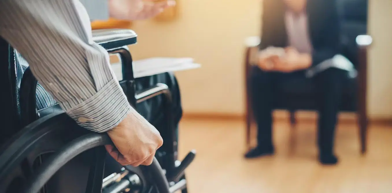 Close-up image of a disabled person's hands gripping a wheelchair wheel, in a formal setting where a man in a business attire is sitting in front of him 