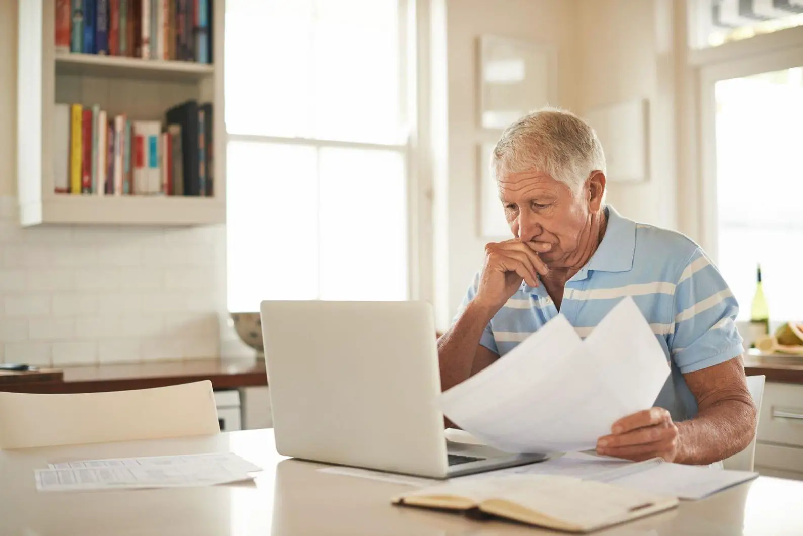 Elderly man diligently working on a laptop, reviewing documents in a seated position.