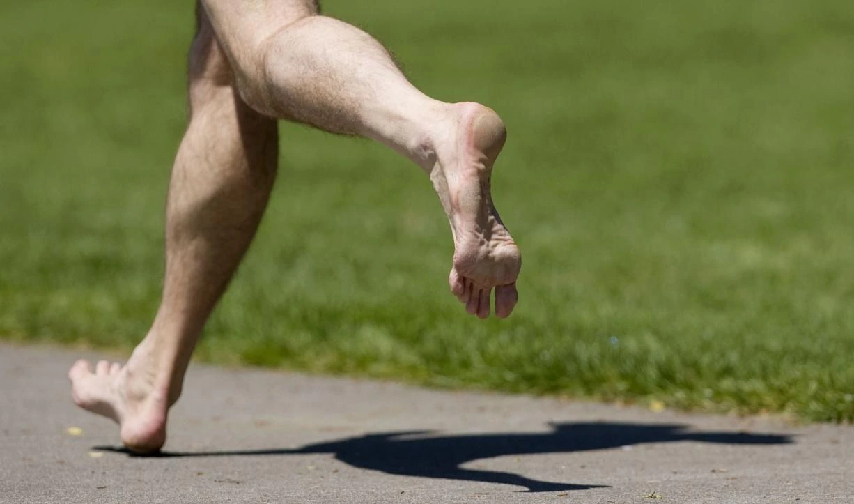 A barefooted man running along a concrete road, with lush greenery on one side. 