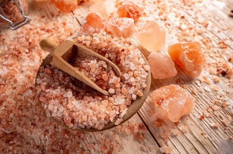 A wooden plate, with pink salt in it, that is placed on a wooden surface, surrounded by scattered large and small salt stones