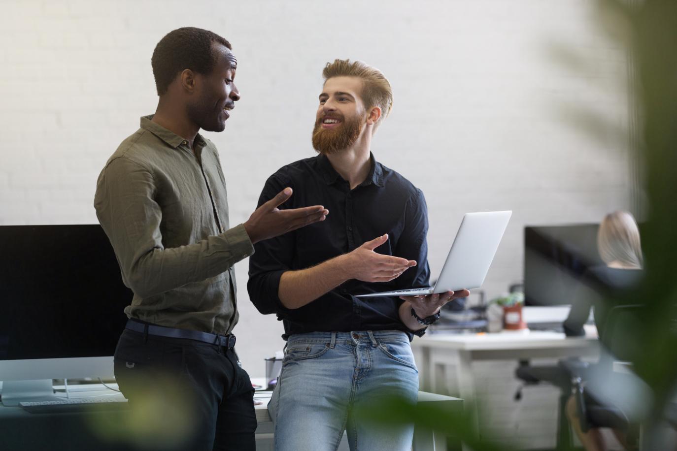 2 Office colleagues talking with each other about their work having an open and purposeful conversation while one is holding a laptop in his one hand, 
