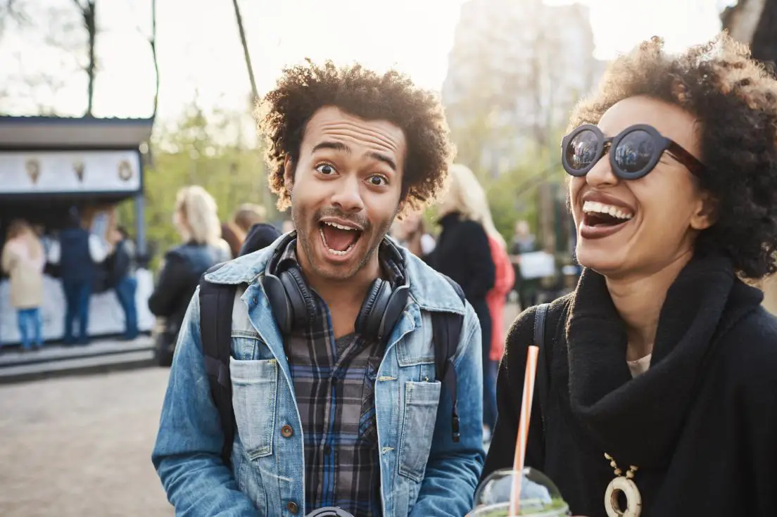 A man and a woman in a part looking in a great mood, both laughing, the boy making a funny face, while girl holding a coffee