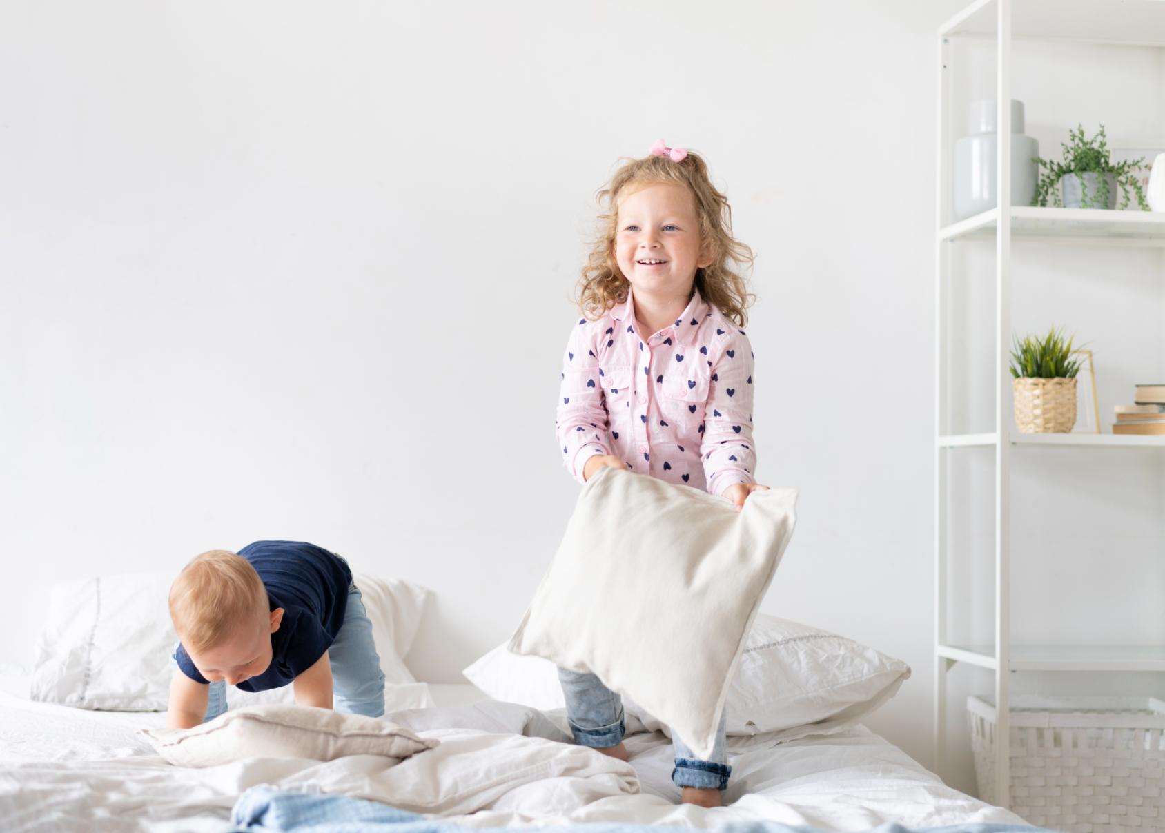 2 little kids on their bed playing with pillows, the girl holding a pillow and smiling