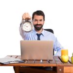 A person sitting in front of his laptop for his official work- showing the clock in his hand with a smile after finising the work on time