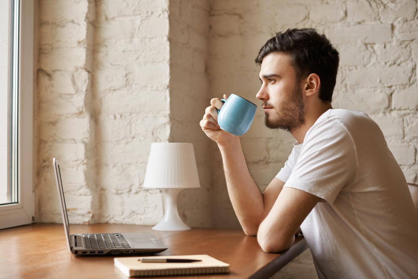 A young man drinking tea while working as his laptop is opened in front of him, trying to reducing his stress from the work
