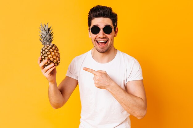 A cheerful man, wearing a white T-shirt and glasses, happily pointing towards a pineapple