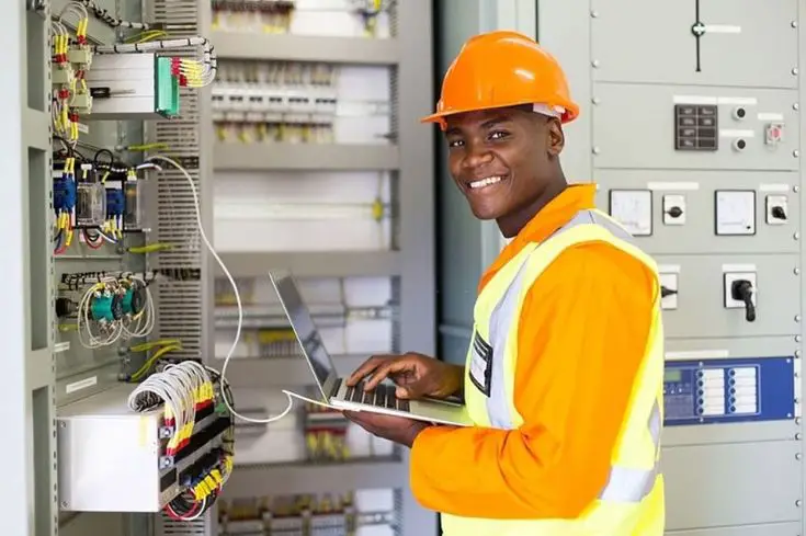 A smiling electrician holding a laptop, standing in front of an intricate integrated circuit board