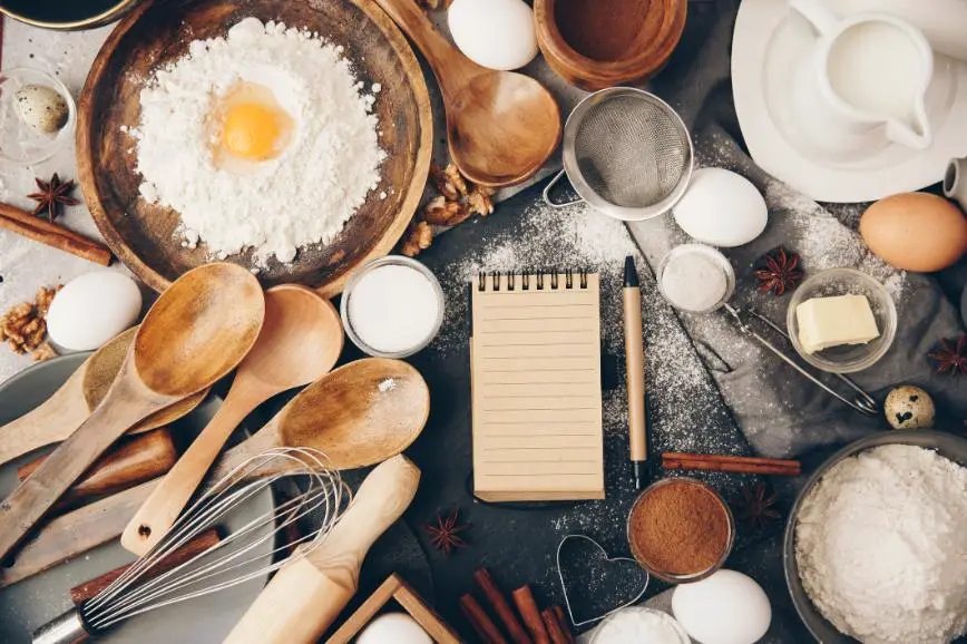 Various kitchen utensils, notepad, flour, eggs, and molds are spread out on a table.
