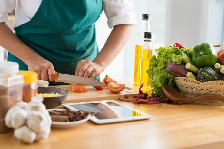 A man wearing an apron cutting tomatoes on a cutting board with various vegetables and spices on the shelf.