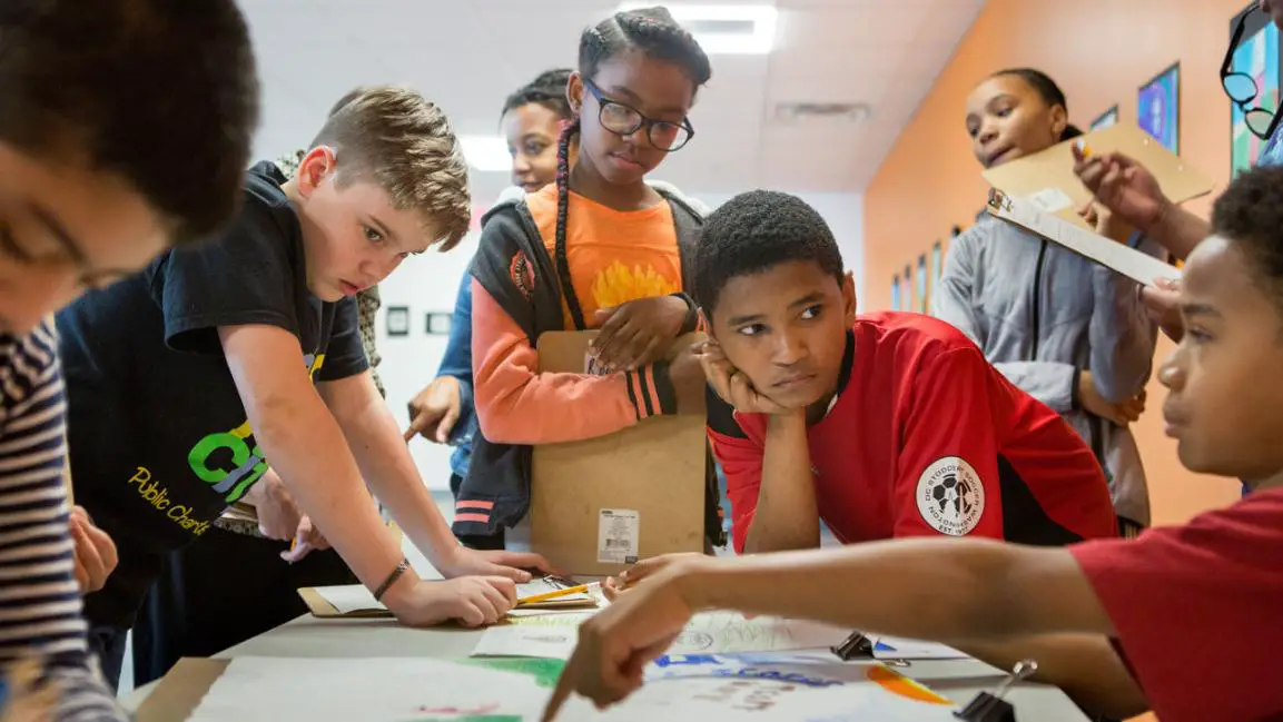 "Children gathered around a table, attentively listening to a black child, explaining them something from the paper