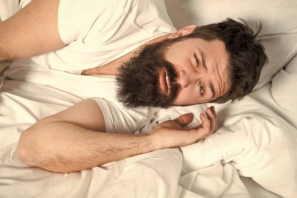 A man on a bed, covered with a white sheet, wearing a facial expression that conveys the struggle with sleep.