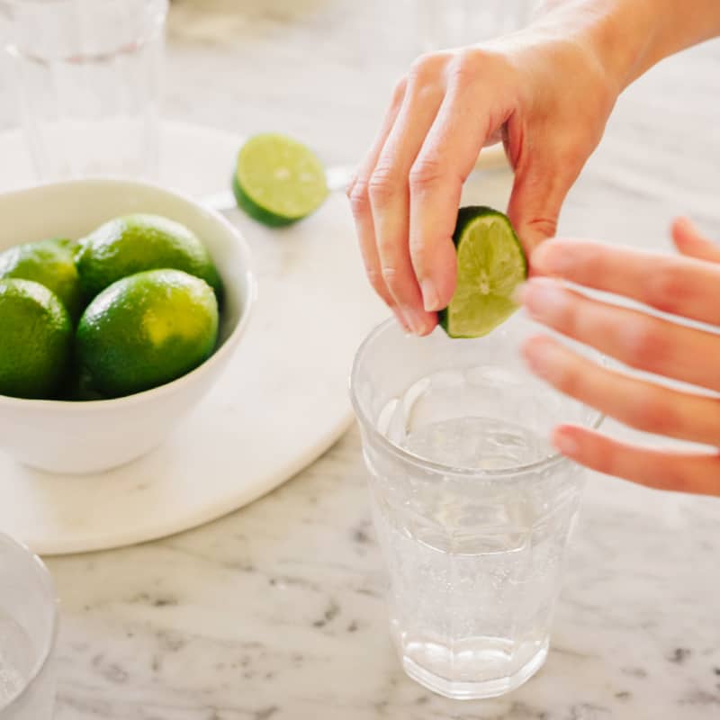 A girl squeezing lime into a glass of water, with a bowl of sliced limes presented on the side.