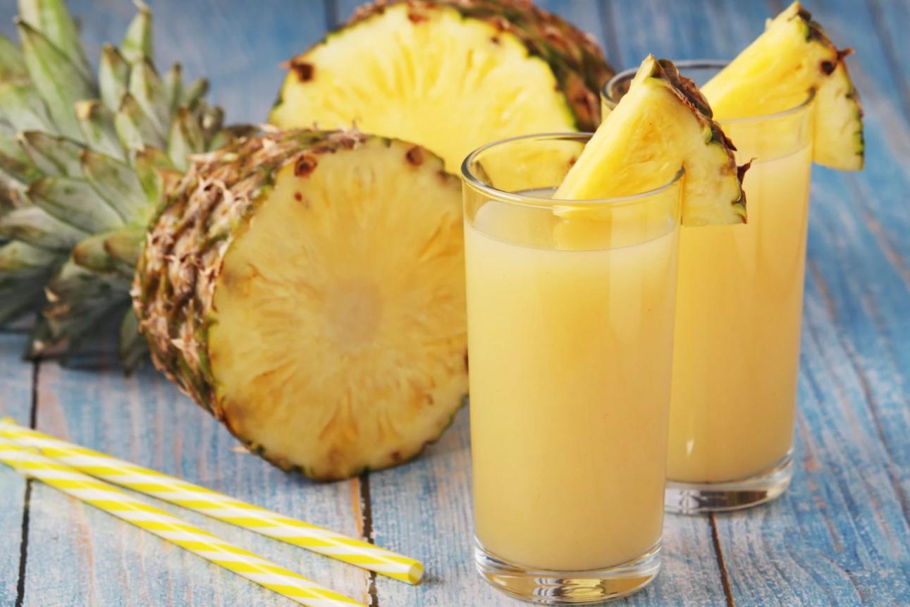 Two glasses of pineapple juice, alomg with a half pineapple and straws, placed on a wooden surface.