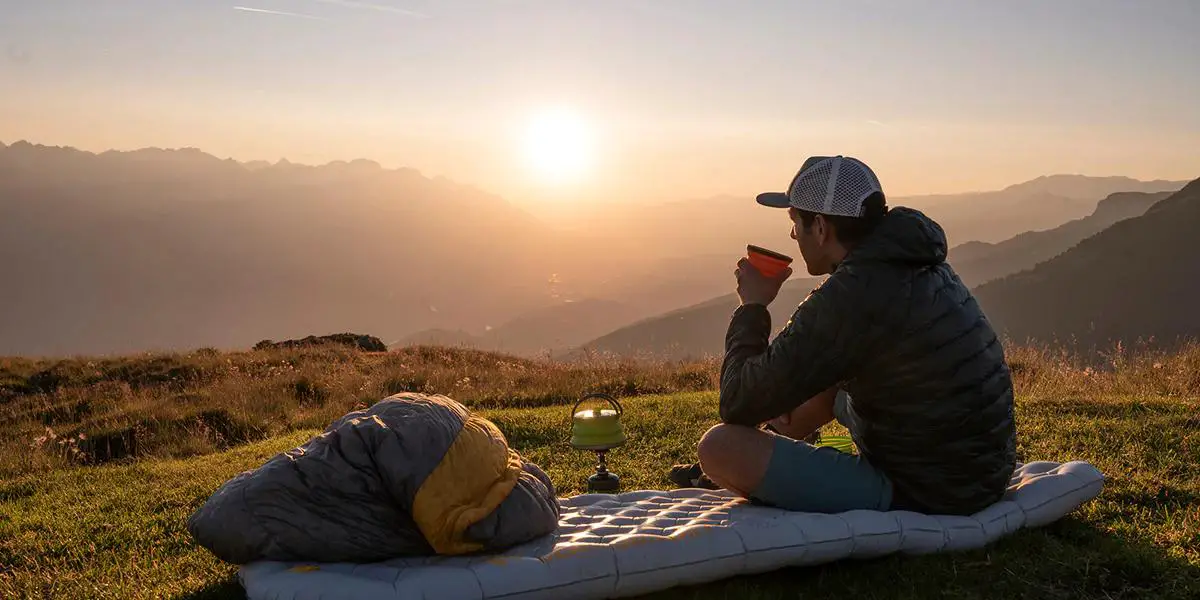 A man sits atop a mountain, comfortably seated on a mat, sipping tea, and gazing at the sun on the horizon