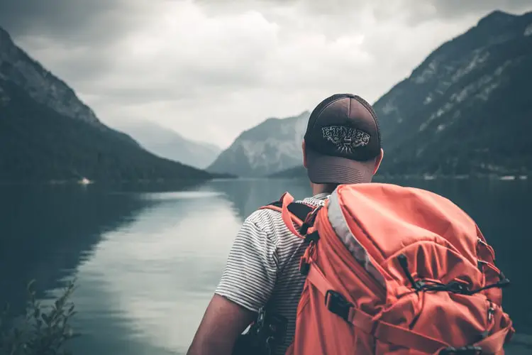 A man standing by a lake with his back turned, wearing a backpack 