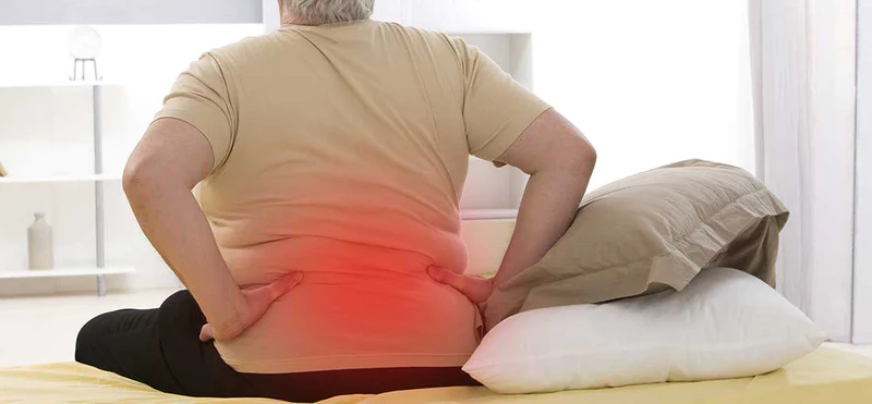 An obese, elderly man with back pain is sitting on a bed