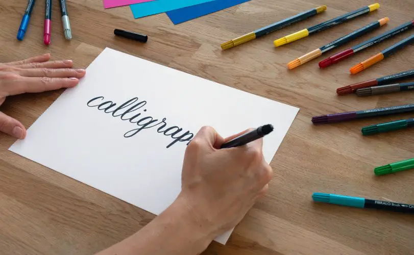 A man writing calligraphy on a paper with various markers arranged alongside on the same table.