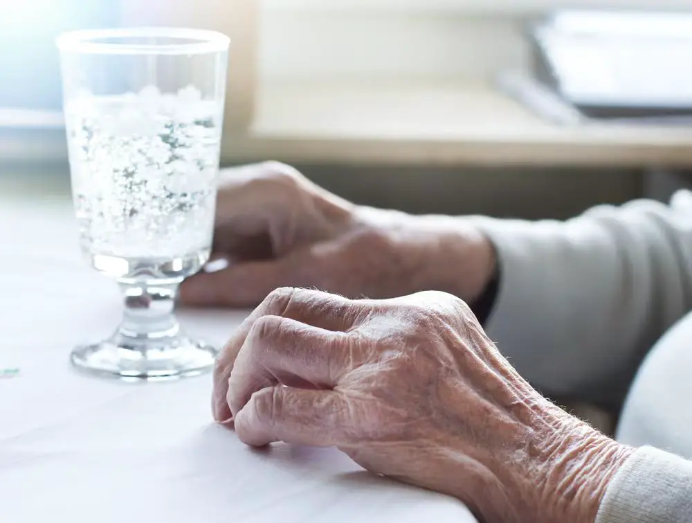 An empty glass resting on a table, accompanied by the weathered hand of an elderly person