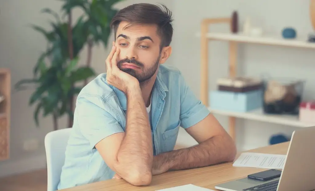 A man, appearing lethargic and despondent, seated at his work table with a laptop in front of him.