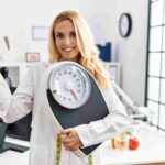 Beautiful blonde nutritionist woman holding weight machine and pointing thumb up towards side