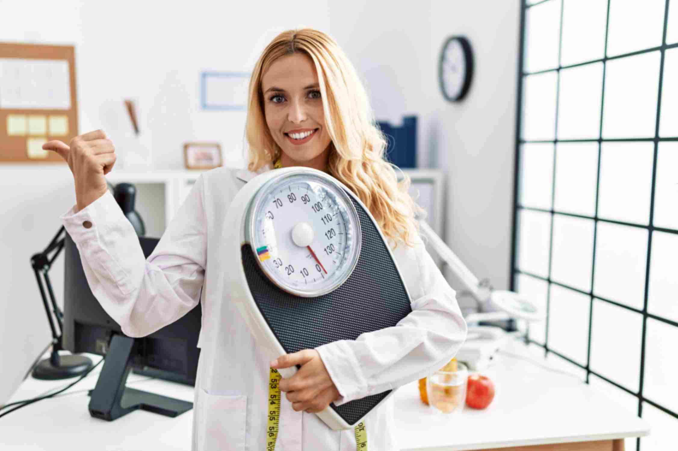Beautiful blonde nutritionist woman holding weight machine and pointing thumb up towards side 