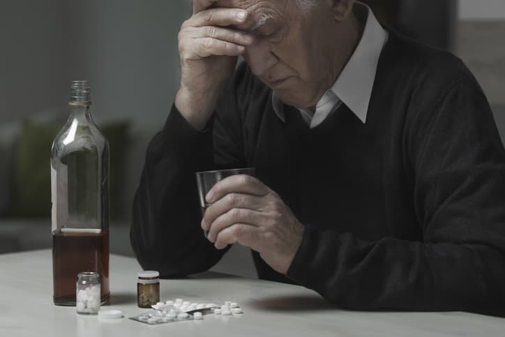An elderly man simultaneously consuming alcohol and pills while holding his head with one hand