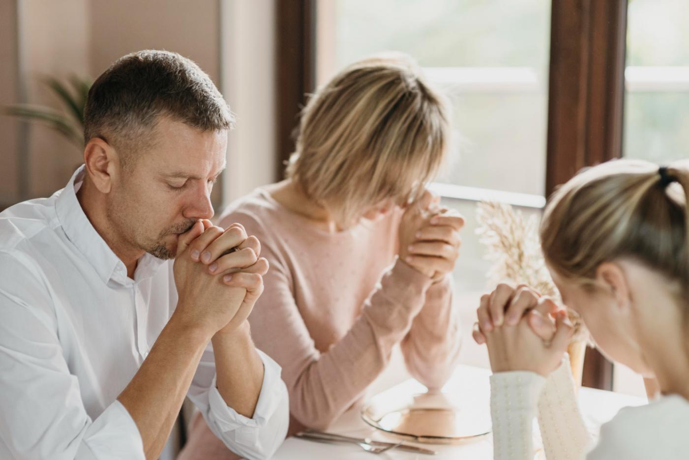3 people from a family with spirituality praying to God sitting on a table in their living room