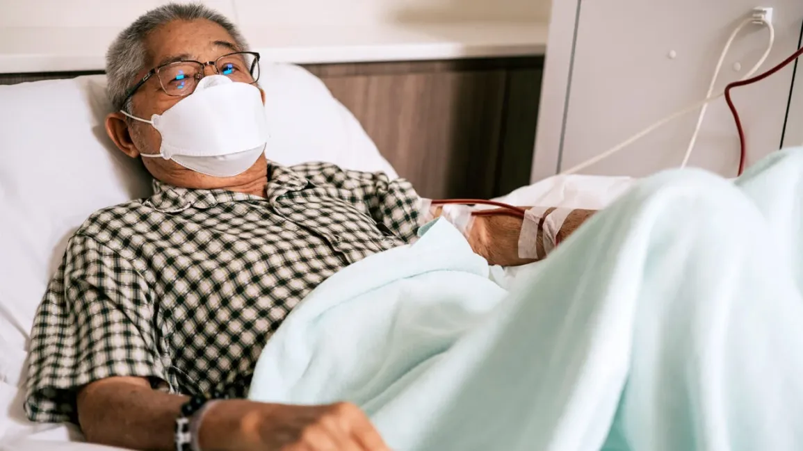 An elderly man wearing mask, dressed in a black and white checkered shirt, is undergoing dialysis in a hospital.