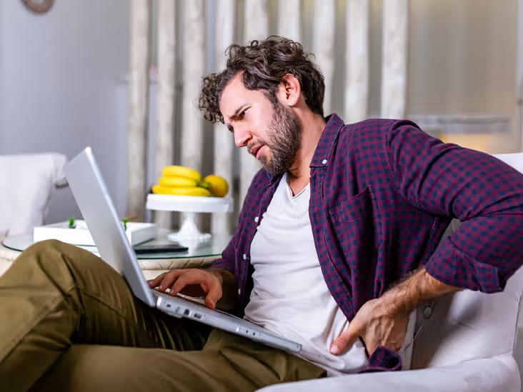 A young man, sitting on a couch with his laptop, displays signs of bad posture, accompanied by evident back pain. 