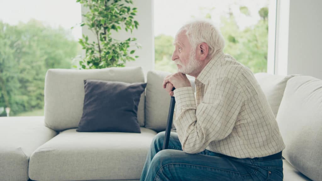 An elderly man sits with a melancholic expression on a sofa, his chin resting on the handle of a walking stick held in both hands