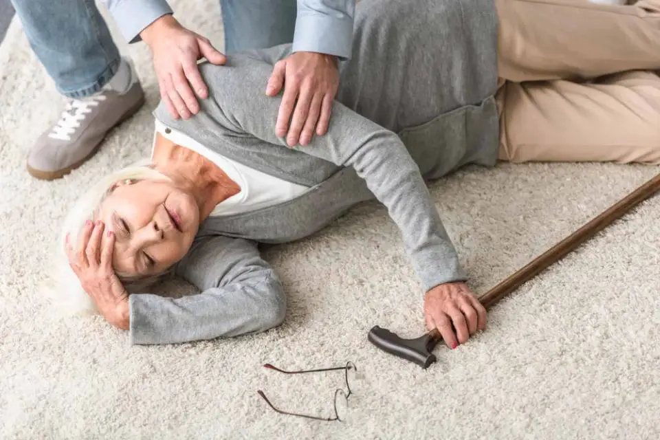 An elderly woman is fallen to the ground, her glasses lying to one side. A concerned man is squatting by her side, ready to assist and offer support.
