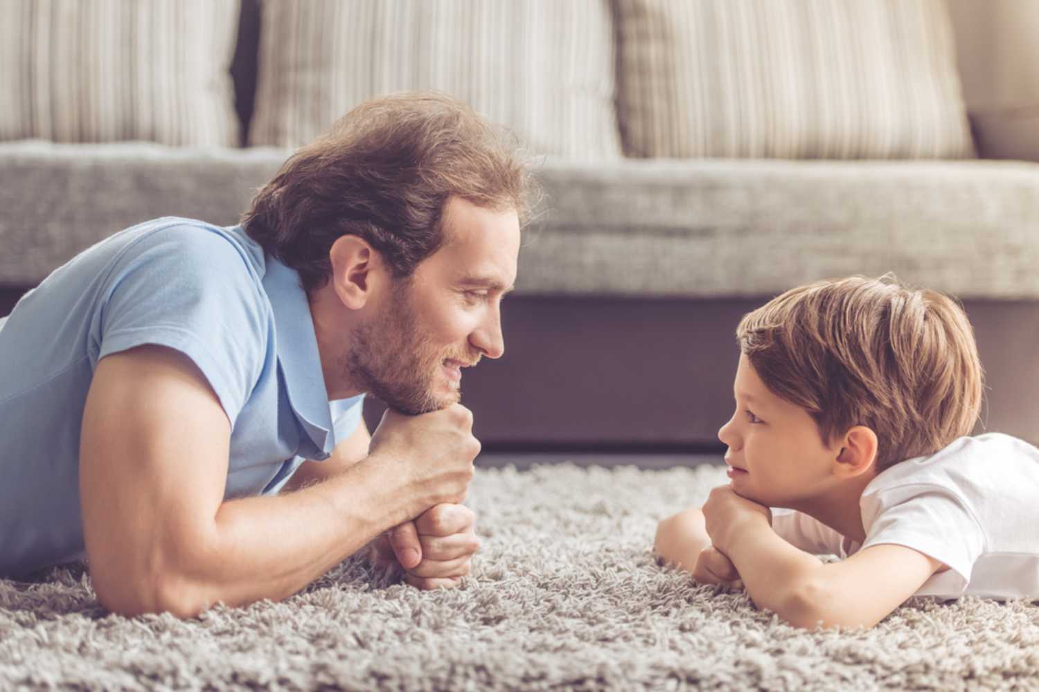 A father and son lying on their stomachs on a rug, resting their heads on their hands, smiling at each other warmly.