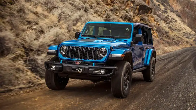 A blue Jeep speeding along a rugged, unpaved road with dry bushes lining one side.