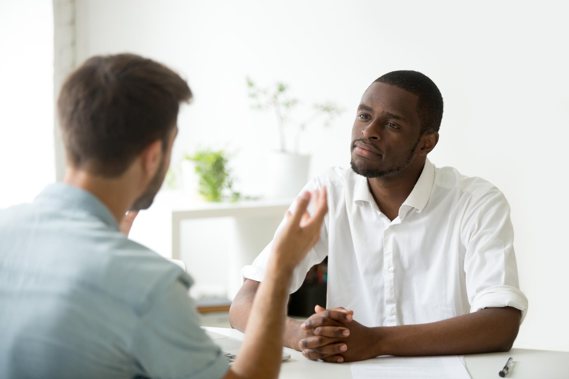 Two individuals engaged in deep conversation: one white man speaking while the other, an black man, listens attentively with direct eye contact.