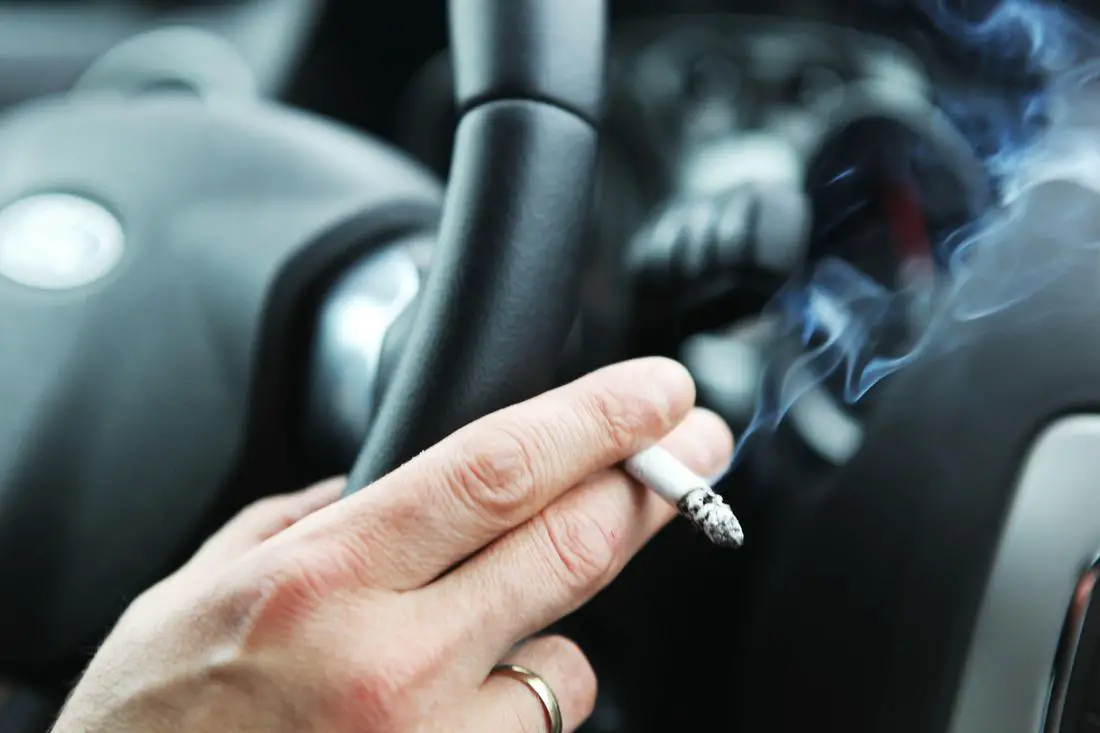 A hand gripping a steering wheel while holding a lit cigarette