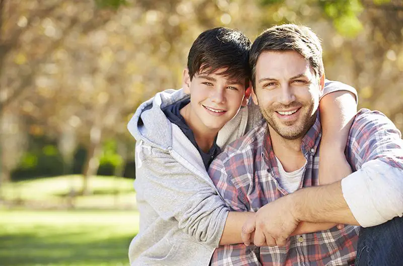 A father and son smiling warmly, with the son's arms wrapped around the father in a loving embrace.