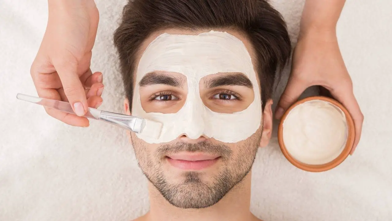 A girl applying a face mask to a man's face using a brush while he lies down.