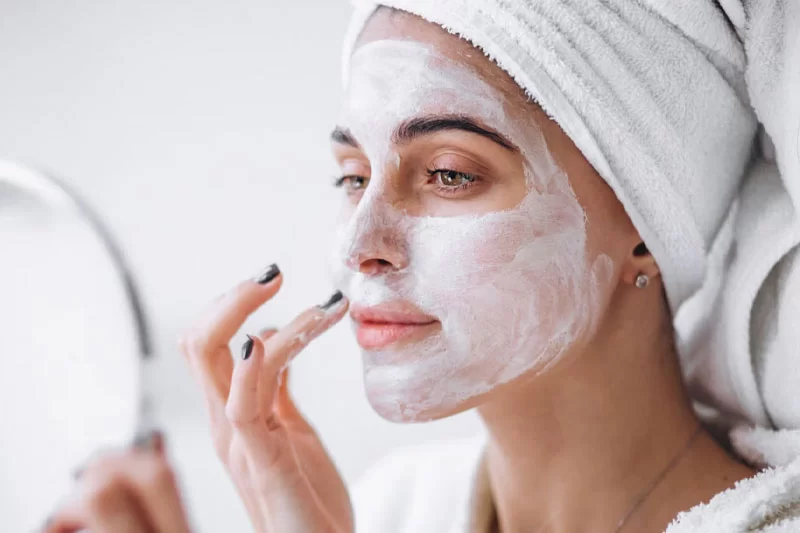 A girl wearing a facial mask with a hair tie wrapped in a towel, admiring her reflection in a handheld mirror.