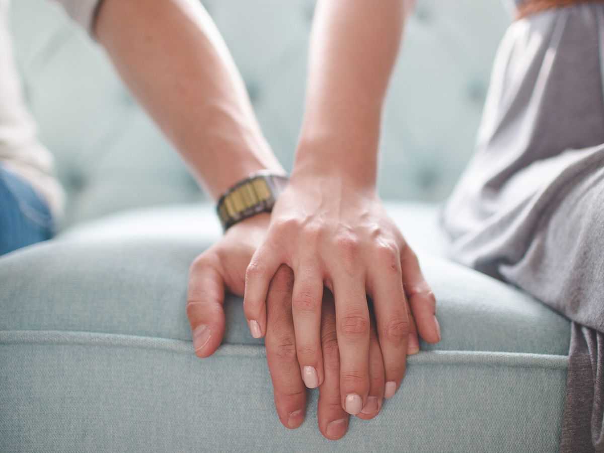 A woman's hand gently rests on a man's hand, conveying support and solidarity as they sit together on a sofa.