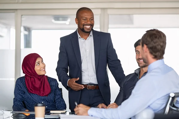 A man laughing and engaging warmly in conversation with two members of his team, while a young woman sits at the same table, observing the interaction.