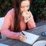 Young woman taking notes outdoors while eating doughnut