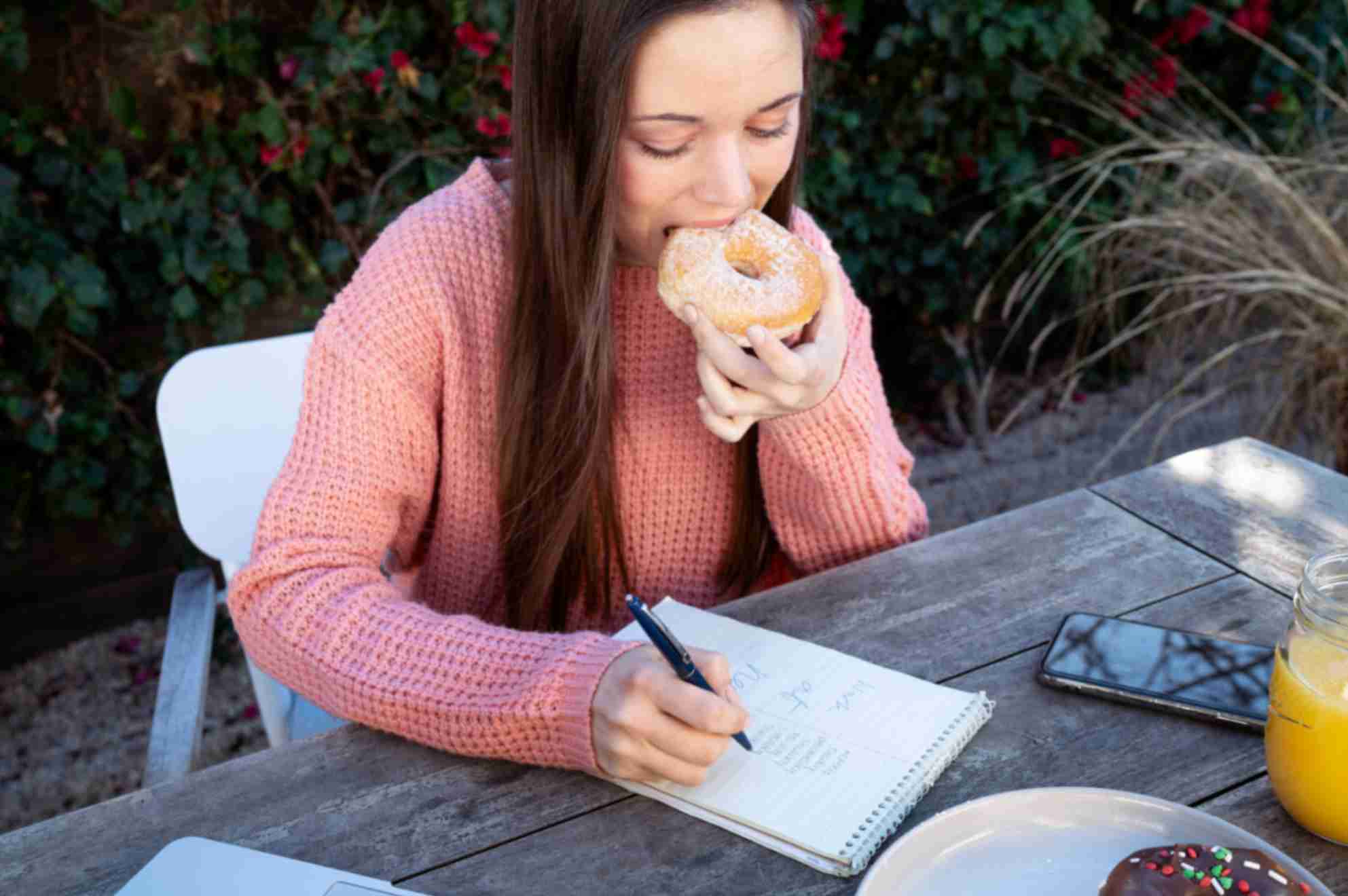 Young woman taking notes outdoors while eating doughnut