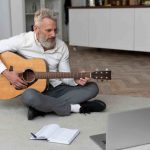 Senior man sitting o home floor taking guitar lessons